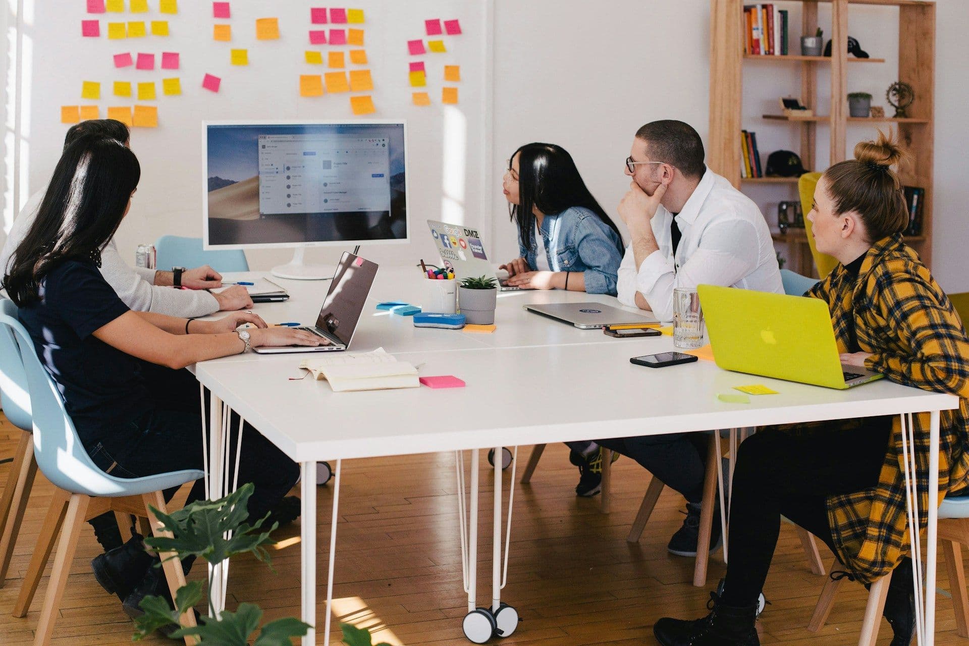 A team meeting around a white table with laptops, in front of a wall of sticky notes.