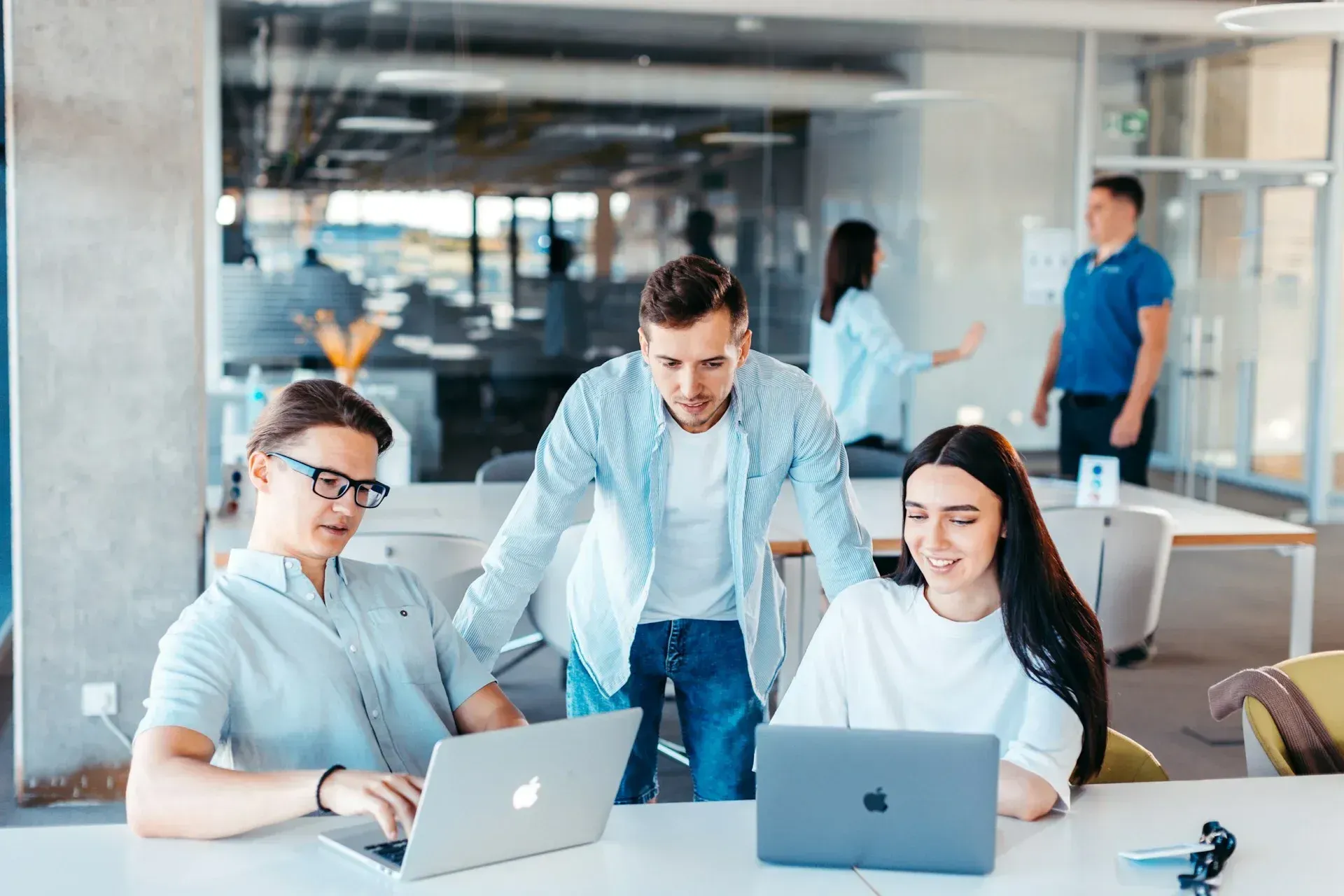 Three people collaborate around laptops in a modern office. Others work in the background.